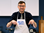 Photo of Adam wearing a white apron with the Braille Bites logo. He is pointing down with index fingers on both hands at ingredients that are out of frame. 