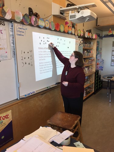 Photo shows a teacher drawing braille characters in print on a whiteboard