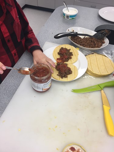 Photo shows a student adding salsa to two tacos on a plate.
