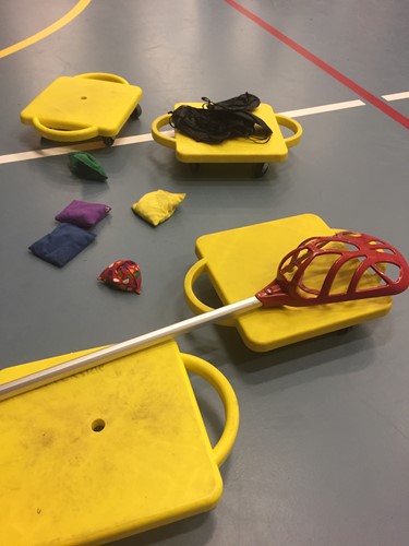 Photo shows scooter boards, bean bags, and a lacrosse stick on a gym floor