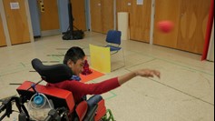 A student using a wheelchair smiles as he throws a ball in a gym.